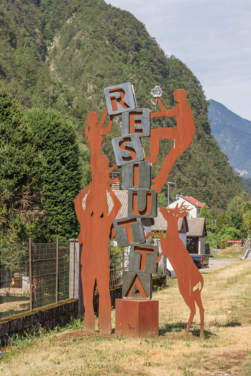 Diese rostfarbene Skulptur mit dem Schriftzug „RESUTTA“ ist eine öffentliche Kunstinstallation am Alpe-Adria-Radweg