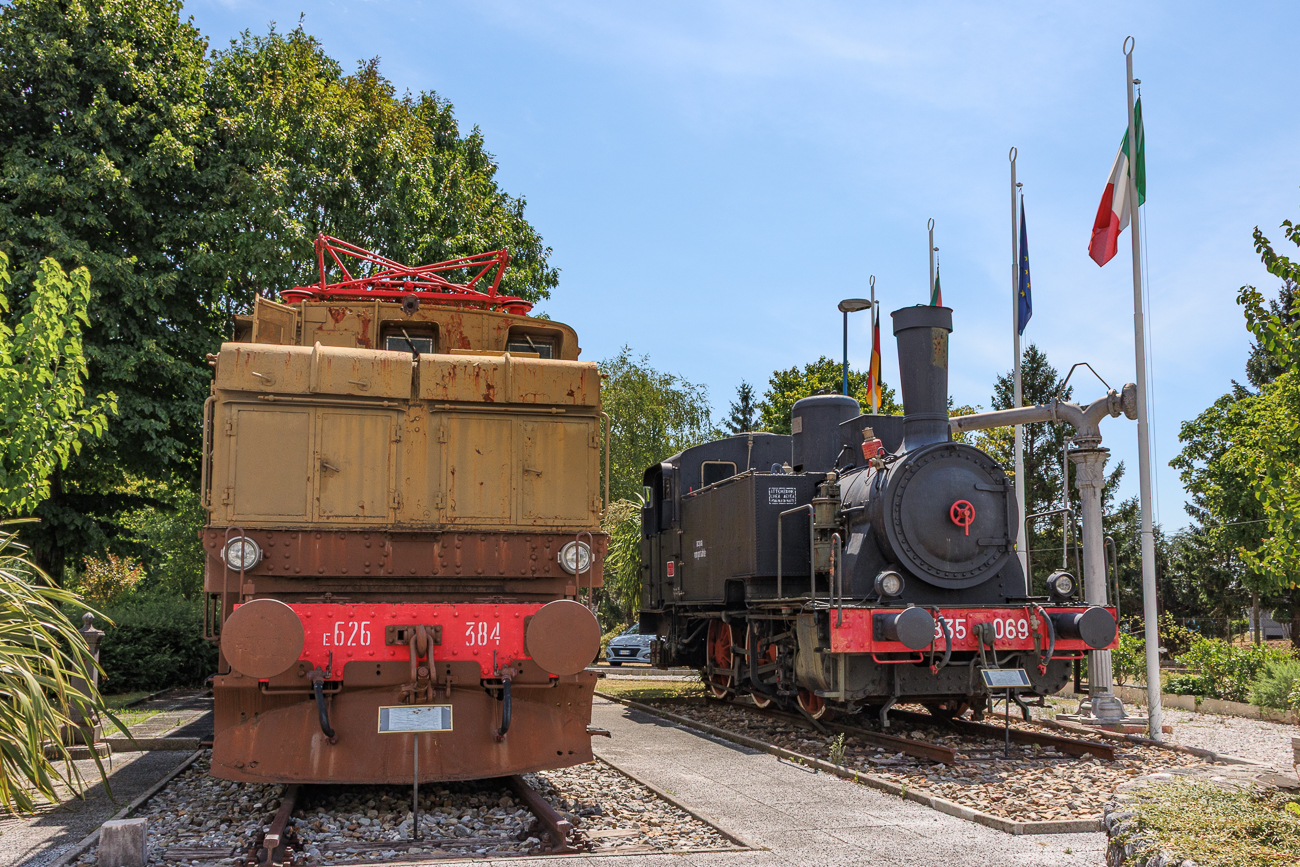 Rechts steht eine historische italienische Tenderlokomotive der Baureihe FS 835 (spezifisch als FS 835.034 erhalten, beschriftet als 835.069). Diese dreifach gekuppelten Rangierlokomotiven pr&auml;gten &uuml;ber Jahrzehnte das Bild auf italienischen Bahnh&ouml;fen.Links daneben ist der markante, braun-beige Vorbau einer alten Elektrolokomotive (E-Lok) der italienischen Staatsbahnen (FS) zu sehen